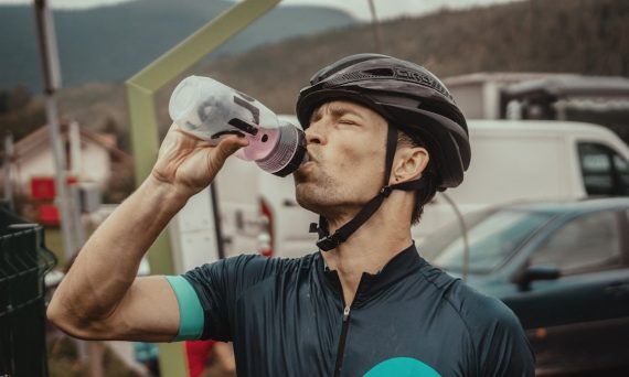man drinking from a sports bottle wearing biking gear in front of parked cars