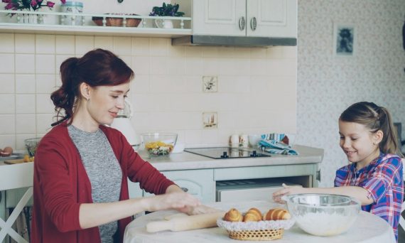 Young mother rolling dough and talking to her little cute daughter while cooking in the kitchen on weekend. Family, food, home and people concept